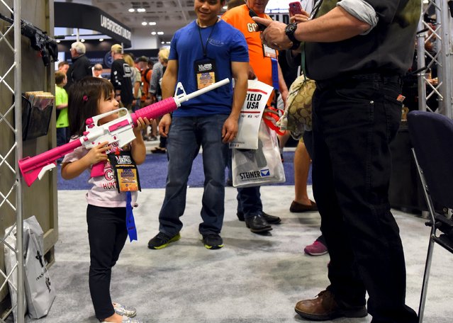 A young girl, who is already an NRA lifetime member, gets her photo made holding a rifle painted pink and white in the trade booths during the National Rifle Association's annual meeting in Nashville, Tennessee April 11, 2015. (Photo by Harrison McClary/Reuters)