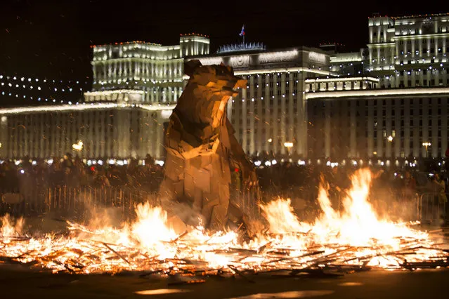 People watch the burning symbol of spring, which looks like a brown bear getting out of his den, created by Hungarian artist Gabor Miklós Szoke for the festival in Gorky Park during a celebration of Maslenitsa, or Shrovetide in Moscow, Russia, Sunday, March 13, 2016, with the building of the National Defense Center of Russian Defense Ministry in the background. Maslenitsa is a traditional Russian holiday marking the end of winter that dates back to pagan times. (Photo by Alexander Zemlianichenko/AP Photo)
