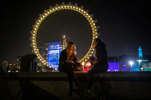 A couple sit with candles on a wall opposite the London Eye which is illuminated yellow on March 23, 2021 in London, England. Marie Curie Cancer Charity has organised a National Day of Reflection on the first anniversary of the coronavirus lockdown for the UK to come together and remember the 126,000 people who have died from Covid-19. A minutes silence will be held at midday and buildings across the country will be lit yellow. (Photo by Rob Pinney/Getty Images)