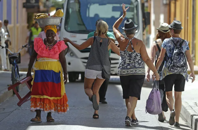 Tourists walk past a “Palenquera”, the name for a woman in traditional costume selling fruit, in Cartagena, Colombia, Friday, October 28, 2016, the city hosting the Ibero-American Summit. “Palenquero” is also the name of a Spanish-based creole language spoken in the area.  (Photo by Fernando Vergara/AP Photo)