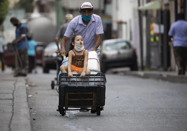 A man, wearing a protective face mask, pushes a dolly filled with empty containers, as he and a child go in search of water in Caracas, Venezuela, Saturday, June 20, 2020, during a relaxation of restrictive measures amid the new coronavirus pandemic. (Photo by Ariana Cubillos/AP Photo)