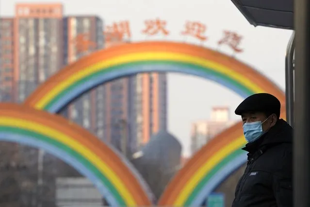 A resident wearing a mask to protect from the coronavirus waits at a bus stop near rainbow decorations on the streets of Beijing on Sunday, December 27, 2020. Beijing has urged residents not to leave the city during the Lunar New Year holiday in February, implementing new restrictions and mass testings after several coronavirus infections last week. (Photo by Ng Han Guan/AP Photo)