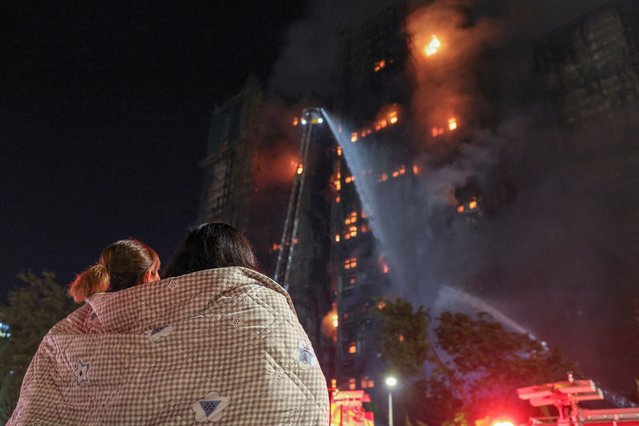 Residents wrapped in blankets watch flames engulf bamboo scaffolding at Wang Fuk Court housing estate after a major fire broke out, in Tai Po, Hong Kong, China, on November 26, 2025. (Photo by Tyrone Siu/Reuters)