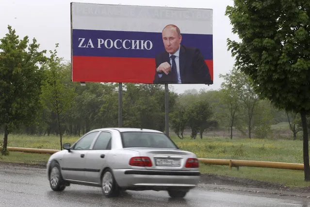 A car drives past a billboard with a portrait of Russian President Vladimir Putin that reads: “For Russia” in Grozny, Russia, Saturday, May 7, 2022. Red Soviet flags and orange-and-black striped military ribbons are on display in Russian cities and towns. Neighborhoods are staging holiday concerts. Flowers are being laid by veterans' groups at monuments to the Great Patriotic War, as World War II is known in the country. At first glance, preparations for Monday's celebration of Victory Day, marking the defeat of Nazi Germany in 1945, seem to be the same as ever. But the mood this year is very different, because Russian troops are fighting and dying again. (Photo by Musa Sadulayev/AP Photo)