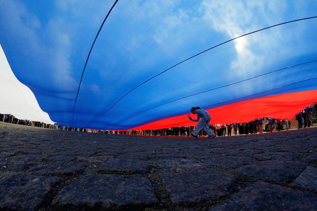 A girl runs under a giant Russian national flag unfurled by volunteers during Russia's National Flag Day celebration in St. Petersburg, Russia, Friday, August 22, 2025. (Photo by Dmitri Lovetsky/AP Photo)