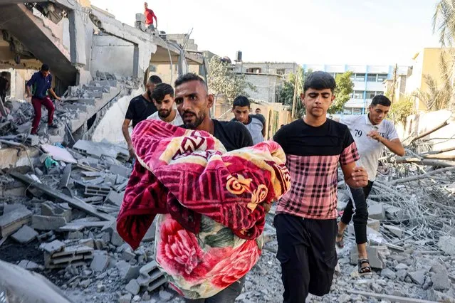 A man carries the remains of an Israeli air strike victim wrapped in a blanket while walking through rubble in Rafah in the southern Gaza Strip on October 13, 2023. Thousands of people – Israelis, Palestinians and foreigners – have been killed since Hamas militants attacked Israel from the Gaza Strip six days ago. Israeli strikes on the coastal enclave have killed more than 1,530 people since Saturday, including 500 children, according to the health ministry in Gaza. (Photo by Said Khatib/AFP Photo)