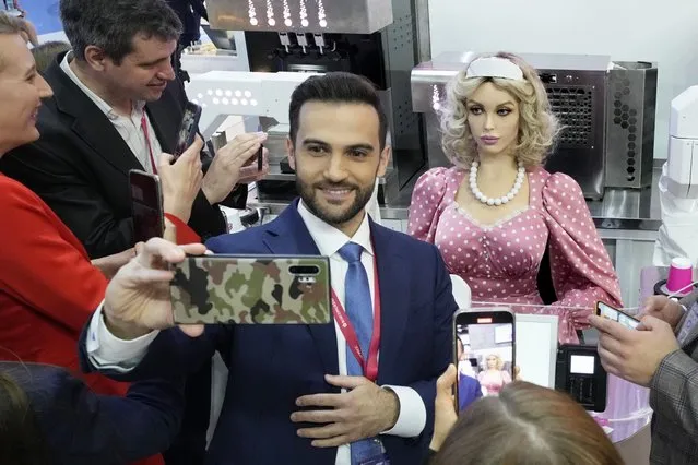 Participants take selfies as Dunyasha, anthropomorphic female cashier robot serves clients on the sidelines of the St. Petersburg International Economic Forum, in St.Petersburg, Russia, Thursday, June 16, 2022. Russia’s annual event to tout its investment opportunities this year was shadowed by the stern international sanctions imposed on the country after the Kremlin sent troops into Ukraine four months earlier and by the extensive disapproval of foreign businesses, which have suspended operations or pulled out entirely, leaving Russian shopping centers pocked with dark, shuttered stores. (Photo by Dmitri Lovetsky/AP Photo)
