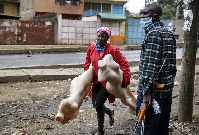 A shopkeeper who sells clothes runs with her mannequins past an armed police officer during clashes between protesters and police in the Kariobangi slum of Nairobi, Kenya Friday, May 8, 2020. Hundreds of protesters blocked one of the capital's major highways with burning tires to protest government demolitions of the homes of more than 7,000 people and the closure of a major food market, causing many to sleep out in the rain and cold because of restrictions on movement due to the coronavirus. (Photo by Brian Inganga/AP Photo)