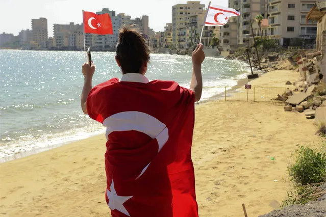 A woman with Turkish and Turkish Cypriot breakaway flags walks along at the beach with abandoned hotels after police open the beachfront of Varosha, an uninhabited, fenced-off suburb in war-divided Cyprus' in the Turkish occupied area in the breakaway Turkish Cypriot north on Thursday, October 8, 2020. The United Nations and the European Union have expressed concern that Turkey's move to open the beachfront could hinder renewed efforts to reunify the island, split in 1974 when Turkey invaded following a coup by supporters of union with Greece. (Photo by Nedim Enginsoy/AP Photo)