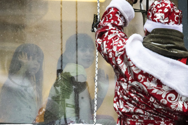 A Russian emergency rescue worker dressed as Ded Moroz (Santa Claus, or Father Frost) greets children as he scales the wall of a children hospital to mark the upcoming New Year celebrations, Russia, Friday, December 25, 2020. Russia, which has so far registered more than 2.9 million confirmed cases of the virus and over 52,000 deaths in the pandemic, has been swept by a rapid resurgence of the outbreak this fall, with numbers of infections and deaths significantly exceeding those reported in the spring. Usually, performers come into children's rooms, but this year because of the virus-related restrictions, the artists had to perform outside of the hospital at a significant distance. (Photo by Pavel Golovkin/AP Photo)