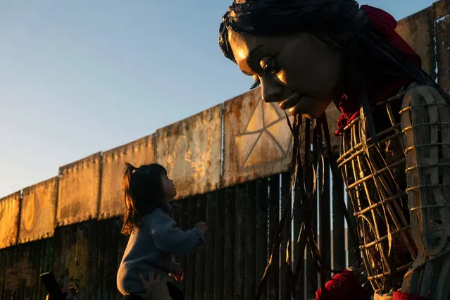 A child greets Little Amal, a 12-foot-tall puppet representing a ten-year-old Syrian refugee child, near the US-Mexico border wall in Playas de Tijuana, Baja California state, Mexico on November 6, 2023. Little Amal is visiting Tijuana within the framework of a journey across Mexico to raise awareness of the plight of refugees and migrants across the globe. (Photo by Guillermo Arias/AFP Photo)