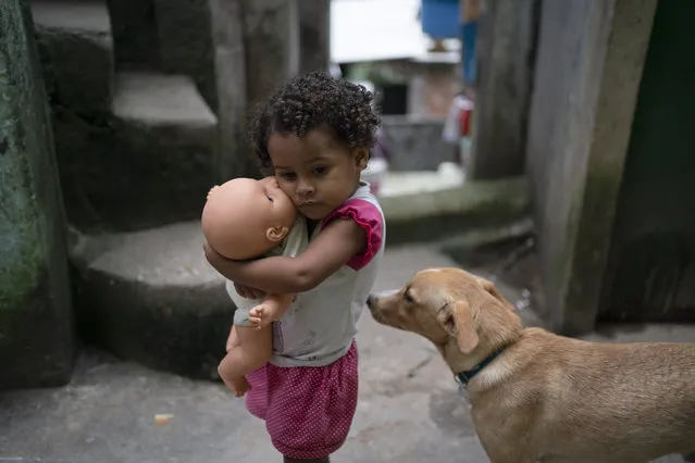 A girl holds her doll in an alley of the Rocinha slum of Rio de Janeiro, Brazil, Tuesday, March 24, 2020. The narrow alleyways of Brazil's largest favela reduce airflow around homes packed tightly together and the poor neighborhood, which lacks proper sewage, has a high incidence of tuberculosis. Authorities are concerned that COVID-19 could easily spread in the favelas of Rio. (Photo by Leo Correa/AP Photo)