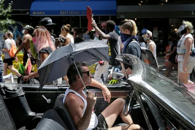 A man in a convertible shields himself from the sun with an umbrella as protesters for social justice walk past in New York, Tuesday, July 28, 2020. (Photo by Seth Wenig/AP Photo)