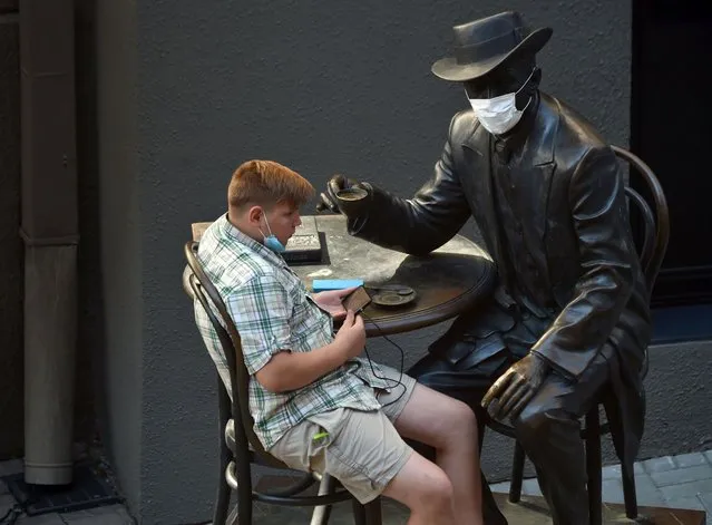 A boy uses his smartphone while sitting on a monument to architect Wladyslaw Horodecki decorated with a face mask during a hot summer day in the Ukrainian capital of Kiev on July 24, 2020. (Photo by Sergei Supinsky/AFP Photo)