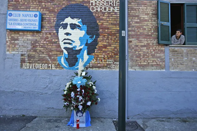 A woman looks out of a window as flowers commemorating soccer legend Diego Maradona are placed beneath a mural depicting him, in Naples, southern Italy, Thursday, November 26, 2020. Maradona died Wednesday, November 25, 2020 in Buenos Aires. (Photo by Alessandra Tarantino/AP Photo)