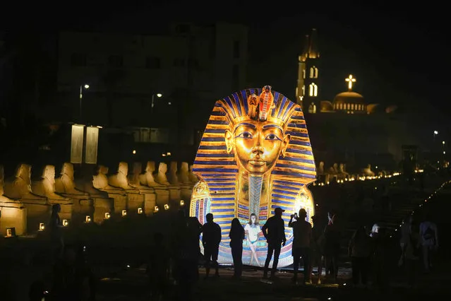 Tourists stand in front of a giant banner showing the golden mask of King Tutankhamun at the Avenue of Sphinxes commonly known as El Kebbash Road in Luxor, Egypt, Friday, November 4, 2022. Egypt celebrates the 100-year anniversary of the discovery of Tutankhamun tomb on Nov. 4, 1922 by British archaeologist Howard Carter and his team. (Photo by Amr Nabil/AP Photo)