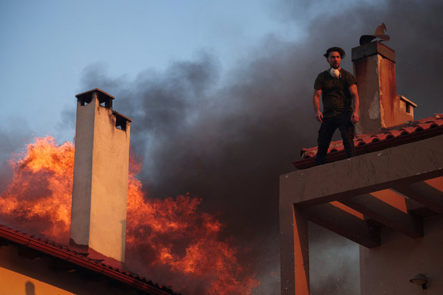A local stands on a rooftop as flames rise from a burning house, while a wildfire burns in the village of Kryoneri, near Athens, Greece on July 26, 2025. (Photo by Alkis Konstantinidis/Reuters)