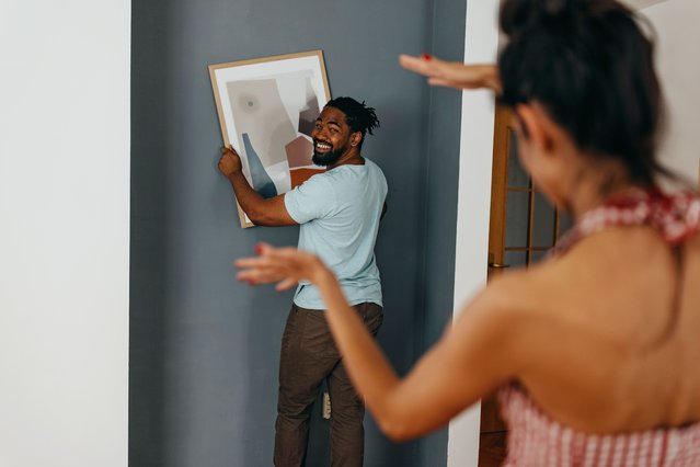 Black man holding framed picture while Caucasian woman is helping to level it against a the wall. (Photo by Getty Images)
