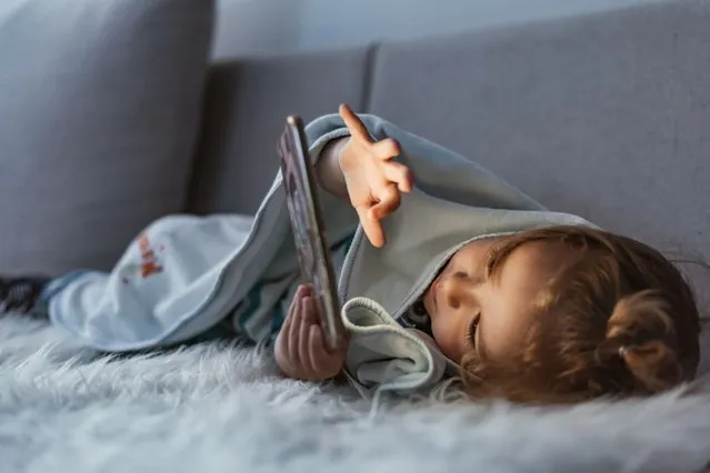 Close up of adorable kid laying down on the bed by holding a smartphone at night. Photo of Little kid in bed under a blanket looking at the smartphone at night. Happy smiling baby boy laying on his side on the bed under a blanket and plays on a smartphone in a game in the dark. The child's face is illuminated by a bright monitor. (Photo by ljubaphoto/Getty Images)