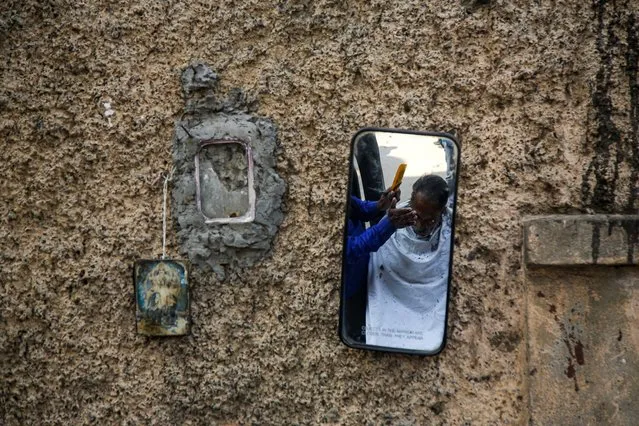 A roadside barber cuts hair, seen in a mirror, beside an image of a Hindu god hanging on a wall, in Kolkata, India, Saturday, January 25, 2020. (Photo by Bikas Das/AP Photo)