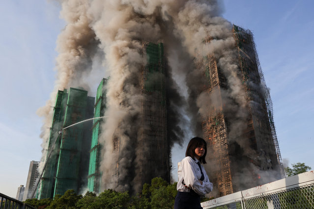 A woman walks as fire and smoke engulf the bamboo scaffolding across multiple buildings at Wang Fuk Court housing estate, in Tai Po, Hong Kong, China, on November 26, 2025. (Photo by Tyrone Siu/Reuters)