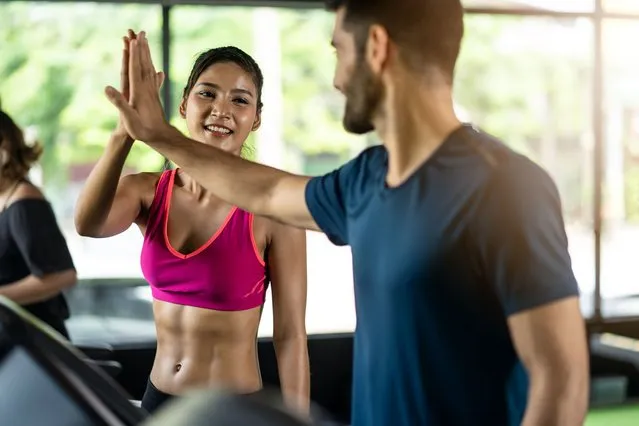 Asian fit beautiful female athlete in sportswear running on treadmill or machine with Caucasian man in fitness gym. Woman raising hand to high five with friend giving encourage, motivation and cheers. (Photo by Kiwis/Getty Images)