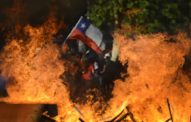 Seen through a burning street barricade, an anti-government demonstrator waves a Chilean flag in Santiago, Chile, Monday, October 28, 2019. Fresh protests and attacks on businesses erupted in Chile Monday despite President Sebastián Piñera's replacement of eight important Cabinet ministers with more centrist figures, and his attempts to assure the country that he had heard calls for greater equality and improved social services. (Photo by Matias Delacroix/AP Photo)