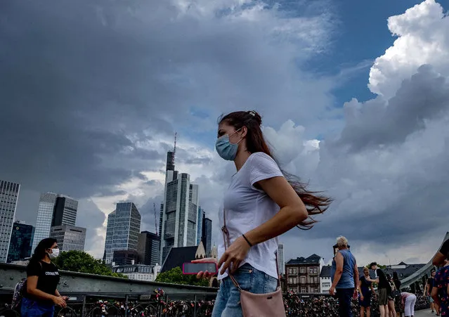 A young woman wears a face mask as she walks on a pedestrian bridge in Frankfurt, Germany, Saturday, August 15, 2020. In background the buildings of the banking district. (Photo by Michael Probst/AP Photo)