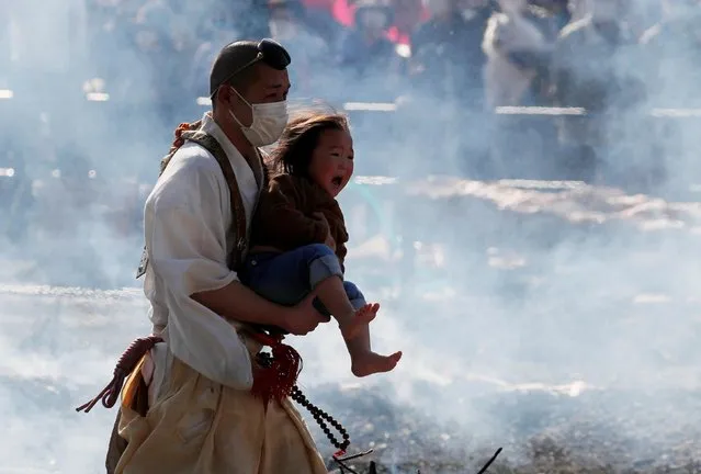 A Buddhist monk wearing a protective mask carries a child as he walks across smouldering coals at the fire-walking festival, called hiwatari matsuri, in Japanese, at Mt.Takao in Tokyo, Japan, March 14, 2021. Japanese worshippers prayed for the safety of themselves and their families by walking barefoot with Buddhist monks over hot coals at an annual festival near Mt. Takaosan. (Photo by Kim Kyung-Hoon/Reuters)