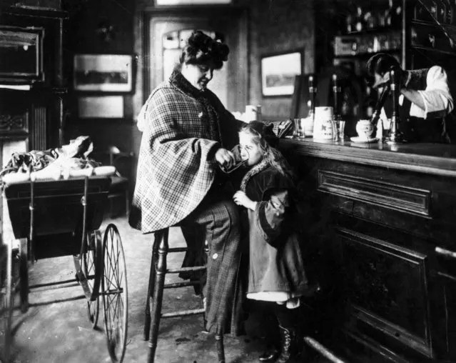 A mother gives her daughter a drink at the bar of a public house, while the baby sleeps in a pram beside her, circa 1900. (Photo by Fox Photos/Getty Images)