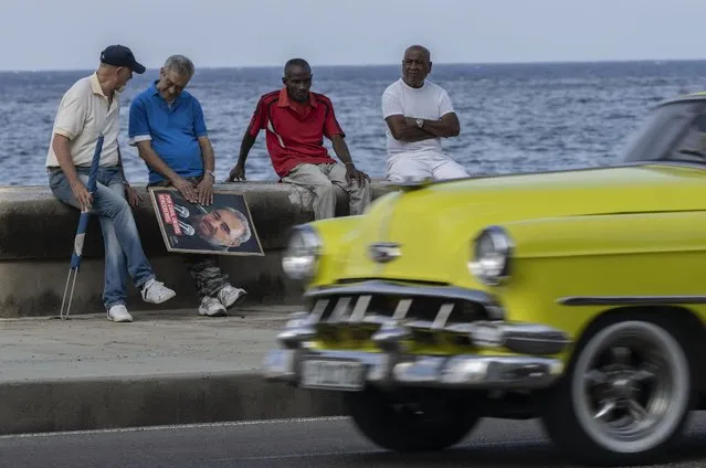 People sit on the malecon with an image of Fidel Castro as they wait for the arrival of the parade of the 65th anniversary of the arrival of Fidel Castro to the capital as head of the rebel Army in Havana, Cuba, Monday, January 8, 2024. (Photo by Ramon Espinosa/AP Photo)