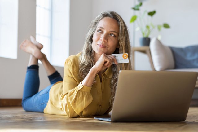 Confident woman at home lying on the floor with laptop holding a cryptocurrency bitcoin credit card looking up. (Photo by Westend61/Getty Images)