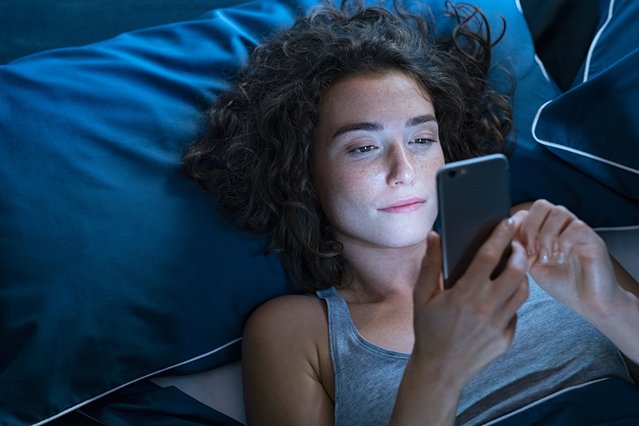 Top view of young woman using a smartphone while lying on bed in the night. High angle view of pretty girl messaging on smart phone before sleeping at night. Addicted woman suffering from insomnia and chatting and surfing on the internet with her cellphone late in night. (Photo by Ridofranz/Getty Images)