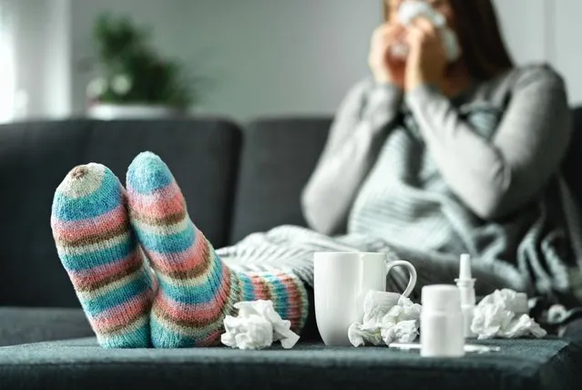Sick woman with flu, cold, fever and cough sitting on couch at home. Ill person blowing nose and sneezing with tissue and handkerchief. Woolen socks and medicine. Infection in winter. Resting on sofa. (Photo by Tero Vesalainen/Getty Images)