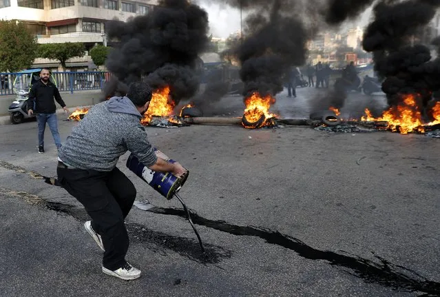 A protester pours oil in front of burning tires blocking a main highway that leads to Beirut's international airport during a protest against the increasing prices of consumer goods and the crash of the local currency in Beirut, Lebanon, Monday, November 29, 2021. (Photo by Hussein Malla/AP Photo)