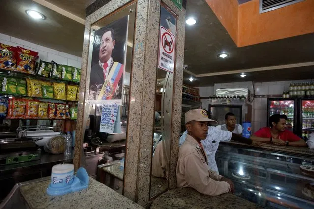 In this March 20, 2017 photo, a militia member stands guard next to a poster of late Venezuelan President Hugo Chavez, at a private bakery that was taken over by the government in Caracas, Venezuela. Within hours of the handover, the new storekeepers took down the Coca-Cola-sponsored sign outside and hung up photos of Maduro, Chavez and South American independence hero Simon Bolivar. (Photo by Fernando Llano/AP Photo)