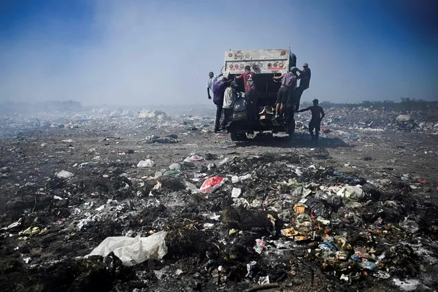 Recyclers hang on to a garbage truck, in the dump of Lujan, Argentina, on December 5, 2022. Dozens of people look for recyclable items among the garbage which they sell to sustain their households and as a way of dealing with the country's economic crisis. The Lujan dump, the largest open-air dump in Argentina, will transform into an environmental center, putting an end to years of pollution, unsanitary conditions, and also to the way in which these recyclers make for a living. (Photo by Luis Robayo/AFP Photo)