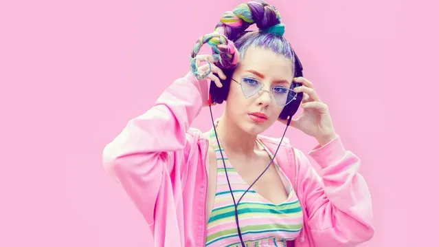 Confident fashionable funky young woman with creative hairstyle wearing cool hoodie standing against pink background and listening music on big headphones while looking at camera. (Photo by Dan Rentea/Getty Images)