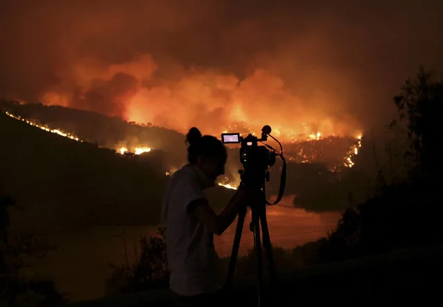A TV camerawoman shoots images of a fire raging up a mountain slope and reflected on the Tagus river outside the village of Sao Jose das Matas, near Macao, central Portugal, Wednesday, July 26 2017. More than 2,300 firefighters with more than 700 vehicles are tackling wildfires in Portugal, where every summer large areas of woodland are scorched. The flames were being driven by powerful winds across steep hillsides of dense pine and eucalyptus forest Wednesday. (Photo by Armando Franca/AP Photo)