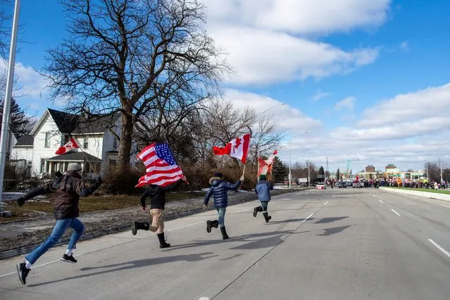 Young adults run with flags towards the protest, as truckers and supporters are being moved back by the police as they continue blocking access to the Ambassador Bridge, which connects Detroit and Windsor, in protest against coronavirus disease (COVID-19) vaccine mandates, in Windsor, Ontario, Canada on February 12, 2022. (Photo by Carlos Osorio/Reuters)