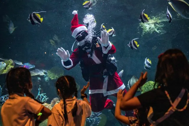 A diver dressed in a Santa Claus costumes waves to young visitors at the Sea Life Bangkok Ocean World aquarium in Bangkok on December 21, 2022. (Photo by Jack Taylor/AFP Photo)