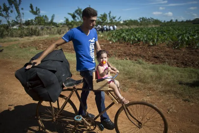 Israel Pardo, 27, pushes a bicycle with his one-year-old daughter while U.S. Secretary of Agriculture Thomas Vilsack speaks to farmers in the background at a rural agricultural cooperative in Guira de Melena, Cuba, November 13, 2015. (Photo by Alexandre Meneghini/Reuters)