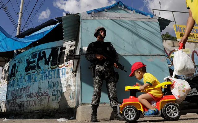 A boy on a cart is pushed near a Brazilian Public-Safety National Force policeman as he patrols an entrance of Chapadao slums complex during a security operation on an effort to crack down on crime in Rio de Janeiro, Brazil, May 15, 2017. (Photo by Ricardo Moraes/Reuters)