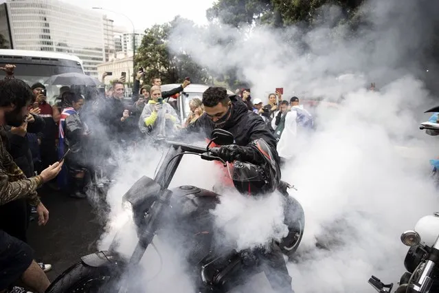Protesters watch as a man spins the tire on his motorcycle in wet conditions as they demonstrate their opposition to coronavirus vaccine mandates at Parliament in Wellington, New Zealand, Saturday, February 12, 2022. The protest began when a convoy of trucks and cars drove to Parliament from around the nation, inspired by protests in Canada. (Photo by George Heard/NZME via AP Photo)