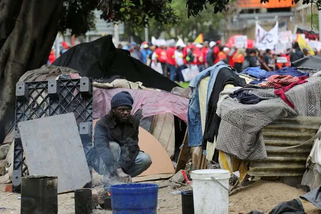 A homeless man prepares food as marchers pass by after the members of South Africa's Congress of South Africa Trade Unions (COSATU), South African Federation of Trade Unions (SAFTU) and other labour unions embarked on a nationwide strike over the high cost of living in Cape Town, South Africa on August 24, 2022. (Photo by Shelley Christians/Reuters)