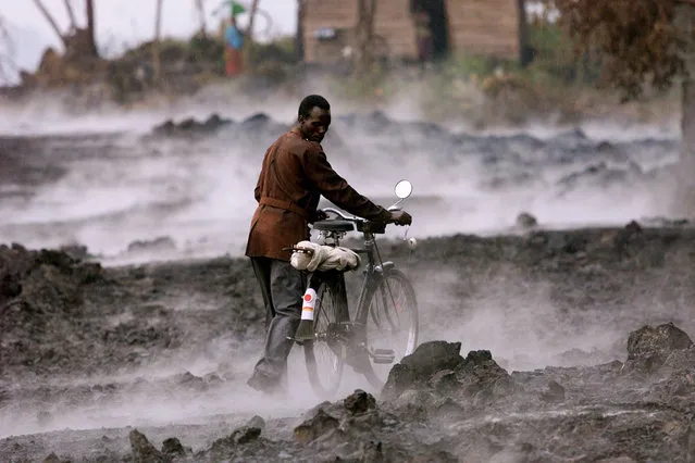 In this file photo taken Thursday, January 24, 2002, a Congolese man pushes his bicycle across lava rock covered in steam, after a rain storm in the eastern Congolese town of Goma. Traumatized farmers are slowly returning to fields decimated by the 2002 eruption of Mount Nyiragongo in eastern Congo. Flowing lava flattened more than 30 percent of the city of Goma, 20 kilometers away. (Photo by Karel Prinsloo/AP Photo)