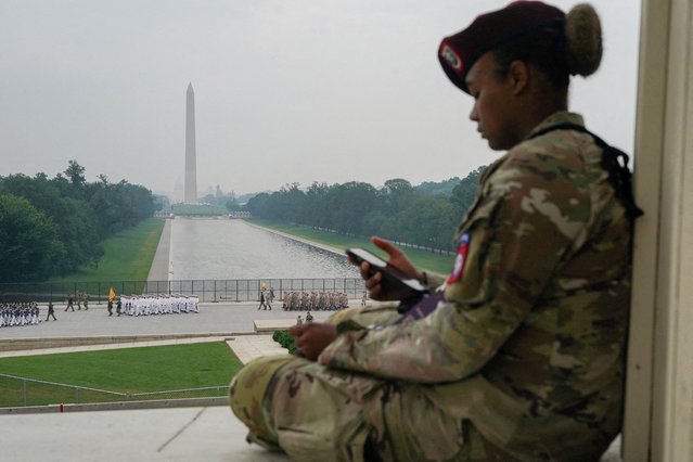 A soldier leans against a pillar while using her phone as other soldiers march with the Washington Monument standing in the background, during a military parade to commemorate the U.S. Army's 250th Birthday in Washington, D.C., U.S., June 14, 2025. (Photo by Nathan Howard/Reuters)