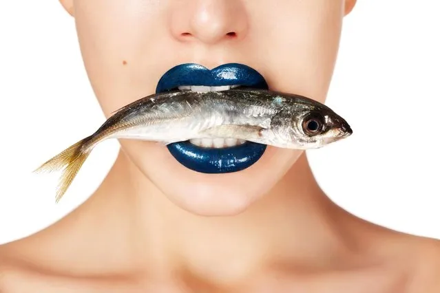 Studio shot of young beautiful woman on light background holding fish in her mouth. Professional make up. (Photo by CoffeeAndMilk/Getty Images)