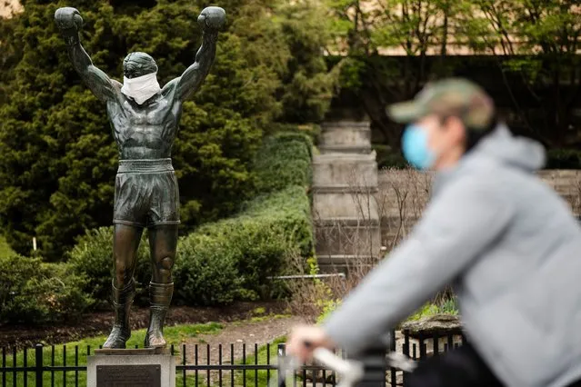 A cyclist wearing a protective face mask as a precaution against the coronavirus moves past the Rocky statue outfitted with mock surgical face mask at the Philadelphia Art Museum in Philadelphia, Tuesday, April 14, 2020. (Photo by Matt Rourke/AP Photo)
