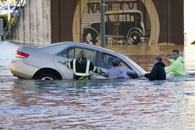 Passersby surround a car whose driver went past a barricade and into the flooded Nooksack River, Tuesday, November 16, 2021, in Ferndale, Wash. About a half-dozen citizens went into the river, where they stopped the car from floating further and muscled it back onshore. No one was injured. An atmospheric river – a huge plume of moisture extending over the Pacific and into Washington and Oregon – caused heavy rainfall in recent days, bringing major flooding in the area. (Photo by Elaine Thompson/AP Photo)
